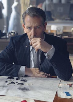 A sombre-looking man sitting at a desk with some fashion designs on the desk in front of him.