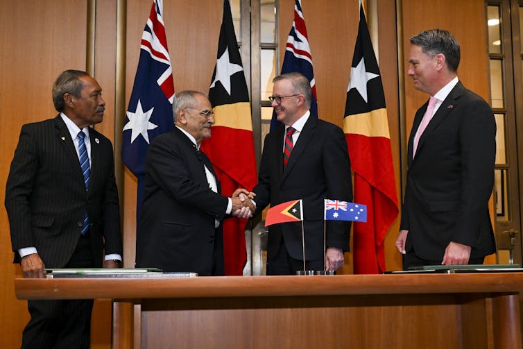 Four middle-aged men in suits stand in an office front of a set of country flags.