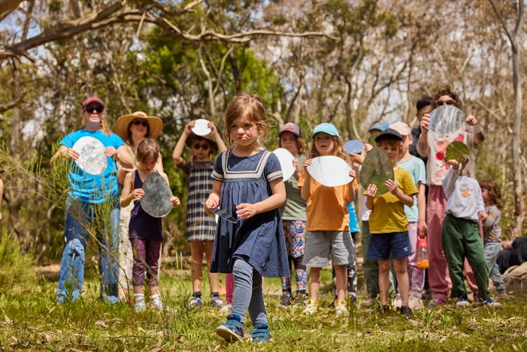 A young girl walks ahead of a crowd of children with mirrors.